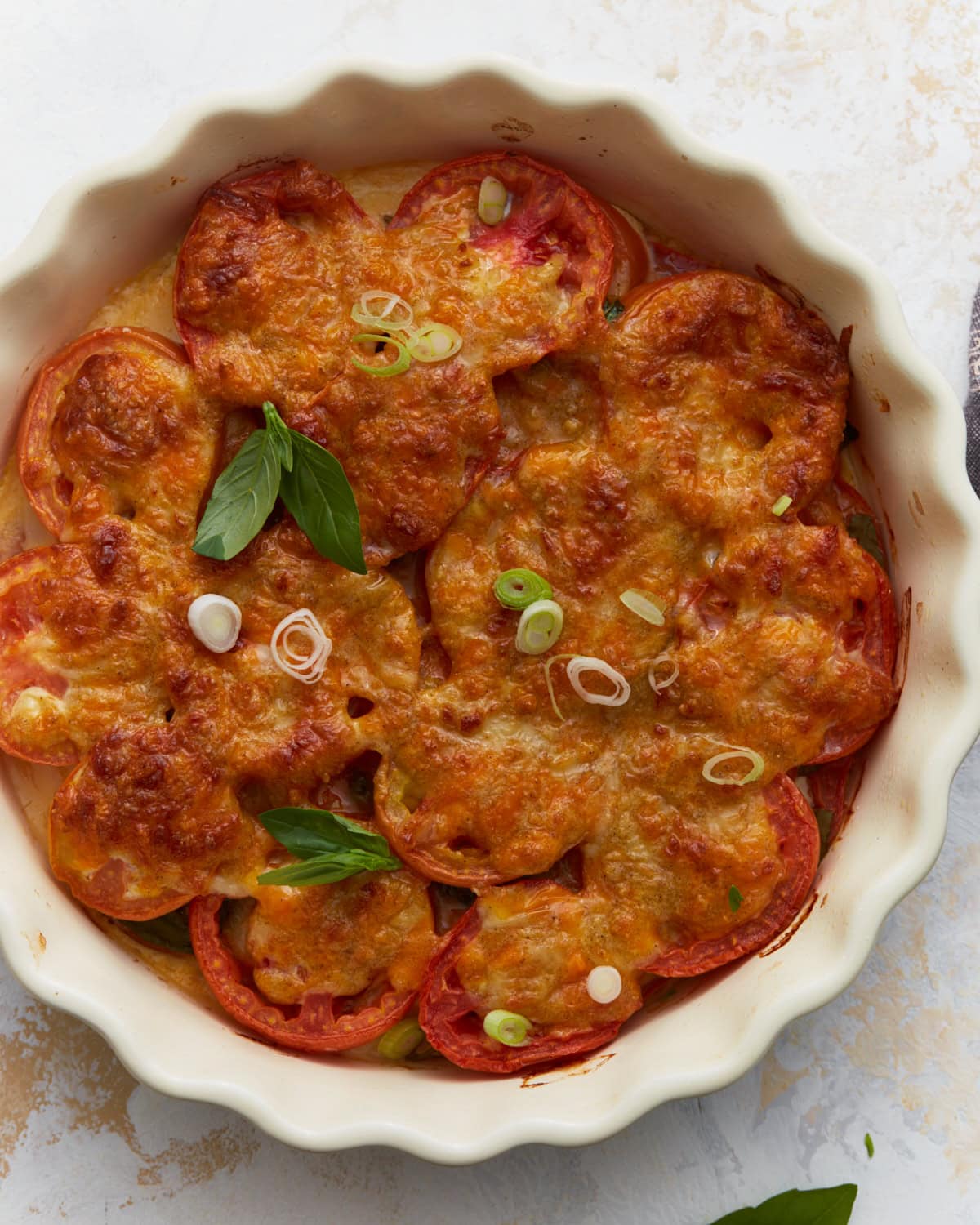 Overhead view of baked crustless tomato pie topped with melted cheese, fresh basil, and sliced green onions in a white pie dish.