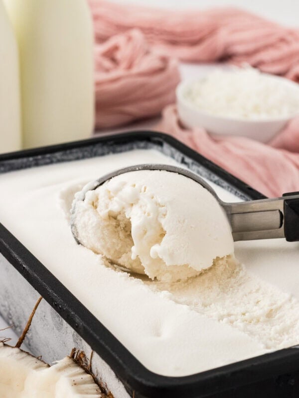 Scoop of creamy coconut sorbet in a loaf pan with shredded coconut and coconut milk in the background.