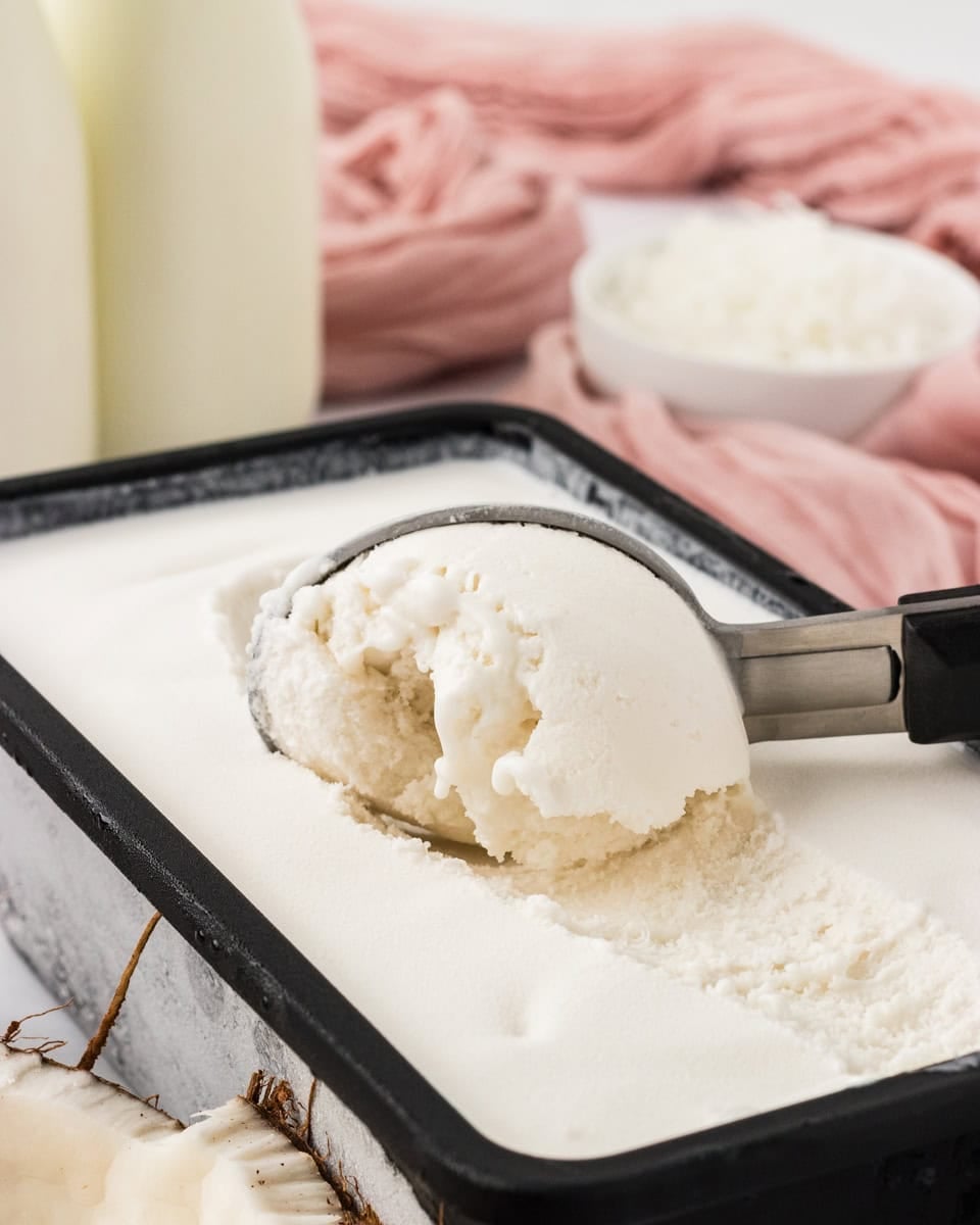 Scoop of creamy coconut sorbet in a loaf pan with shredded coconut and coconut milk in the background.