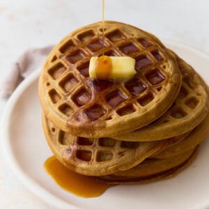 Stack of oat flour waffles topped with butter and maple syrup on a white plate.