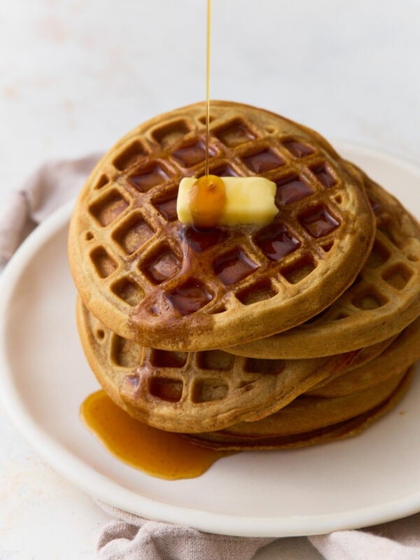 Stack of oat flour waffles topped with butter and maple syrup on a white plate.
