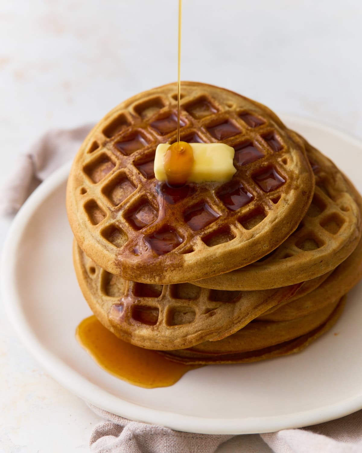 Stack of oat flour waffles topped with butter and maple syrup on a white plate.