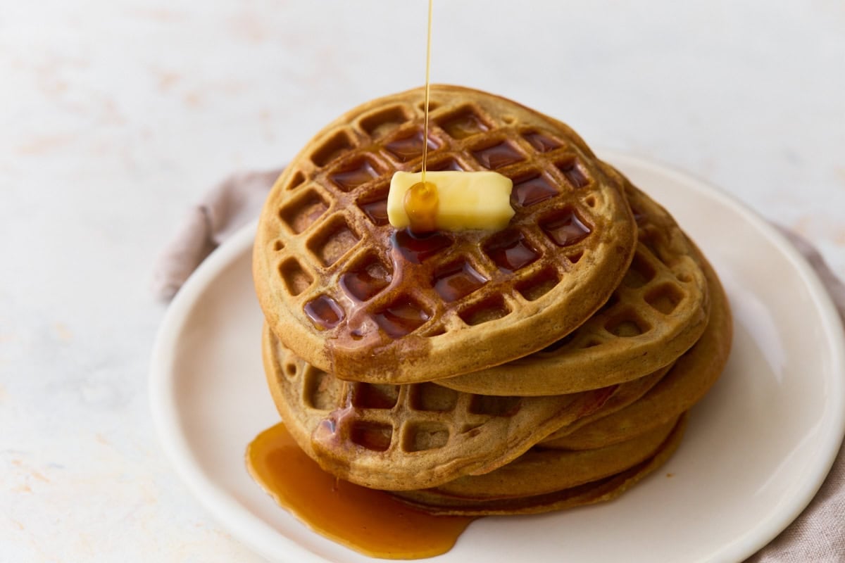 Maple syrup poured over a stack of homemade oat flour waffles with butter.