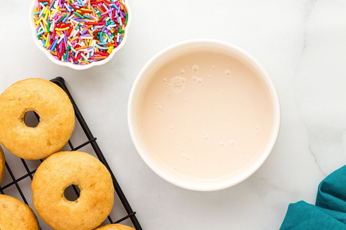 Vanilla glaze in a white bowl beside baked gluten-free donuts and rainbow sprinkles