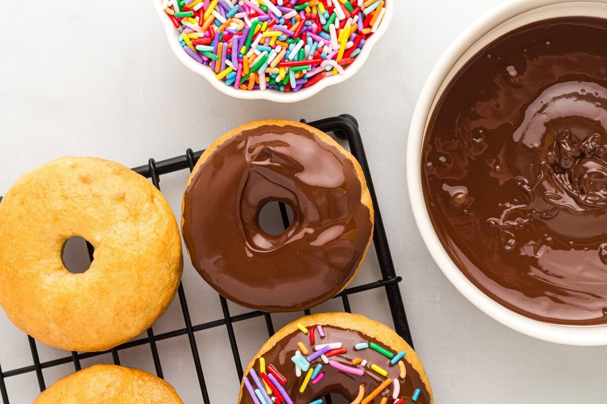 Gluten-free vanilla cake donuts with glossy chocolate glaze beside melted chocolate and rainbow sprinkles.