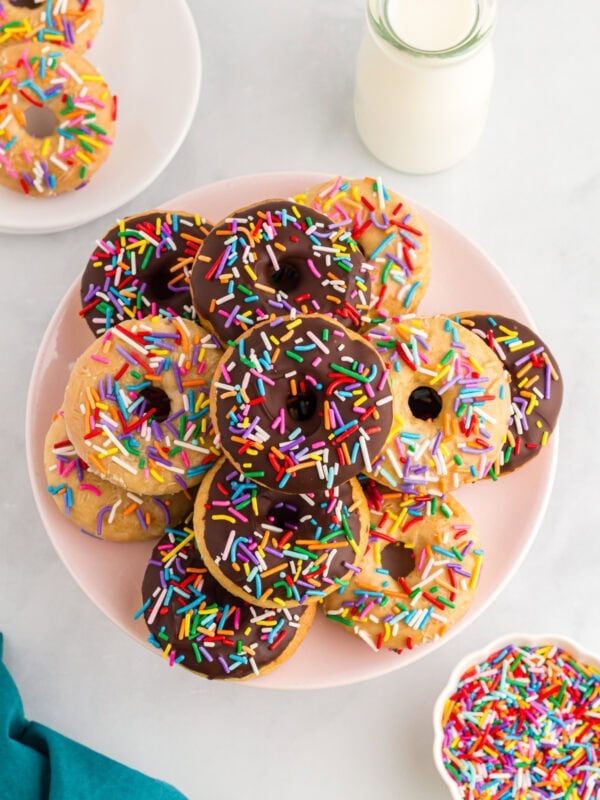 Gluten-free vanilla and chocolate cake donuts topped with rainbow sprinkles on a pink plate beside a bottle of milk.