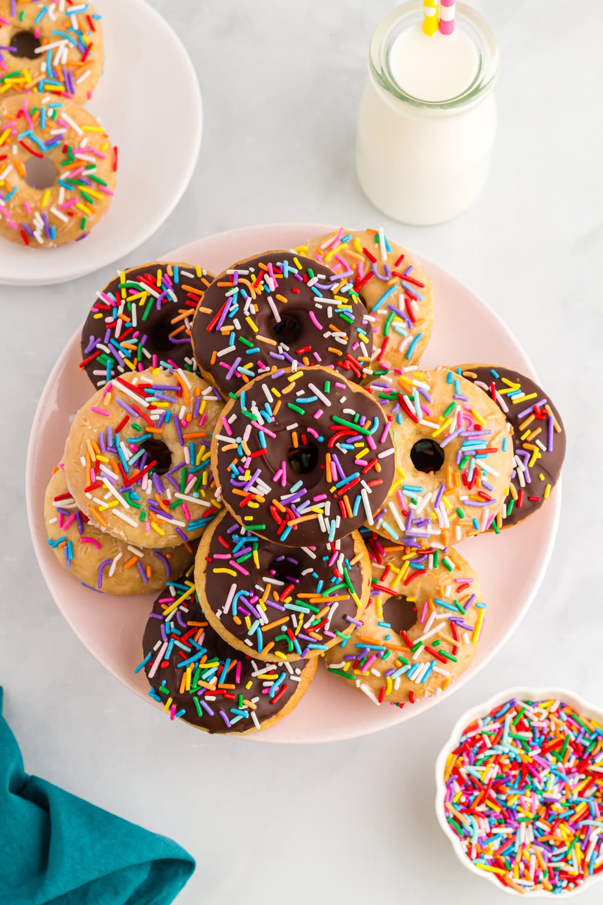 Gluten-free vanilla and chocolate cake donuts topped with rainbow sprinkles on a pink plate beside a bottle of milk.