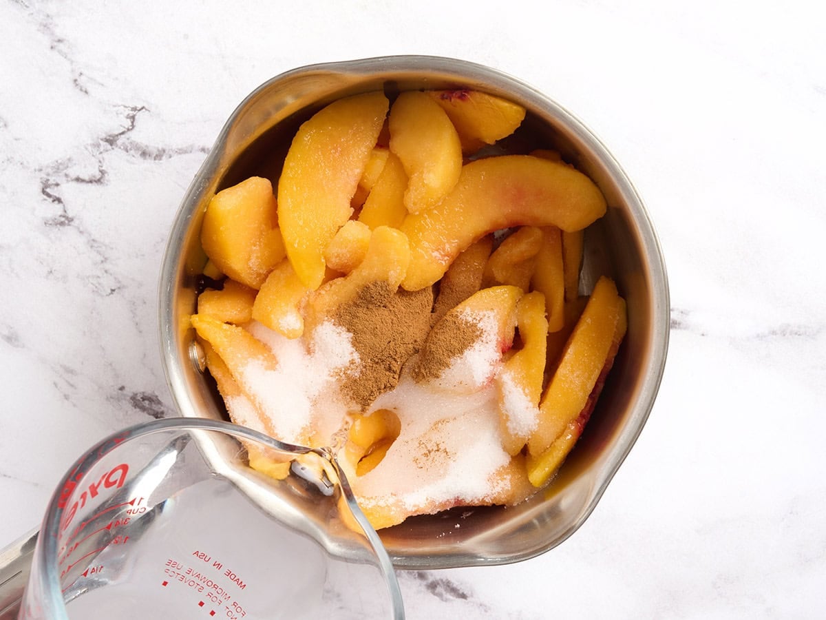 Frozen peach slices, sugar, cinnamon, and water being combined in a stainless steel saucepan
