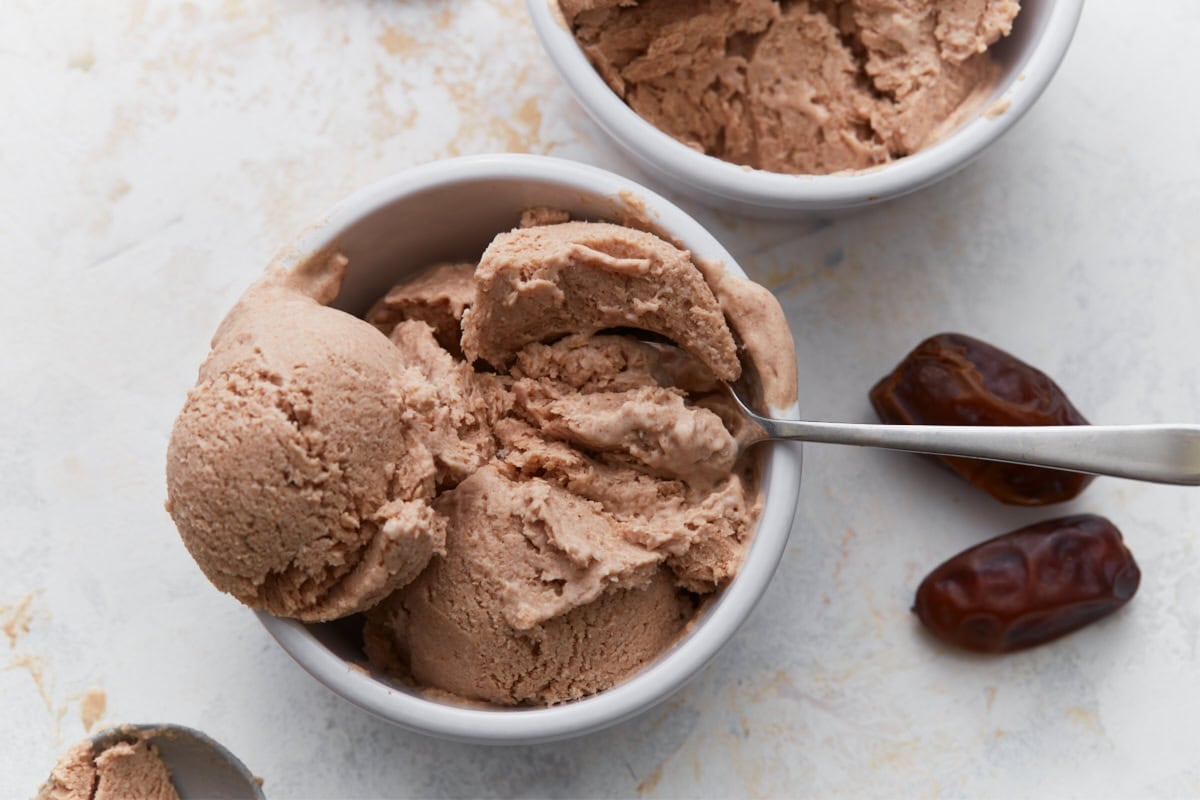 Creamy date ice cream scooped into bowls with whole dates and a spoon on a light background.