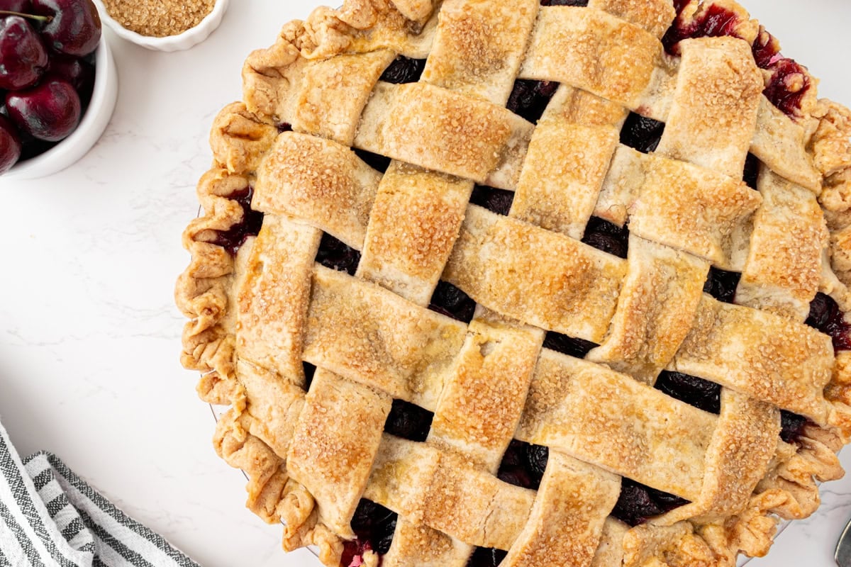 Overhead close-up of a fully baked gluten-free cherry pie with a deep golden lattice crust sparkling with turbinado sugar, a crimped edge, and glossy cherry filling bubbling through the gaps, with a bowl of fresh cherries and a small dish of sugar in the background.