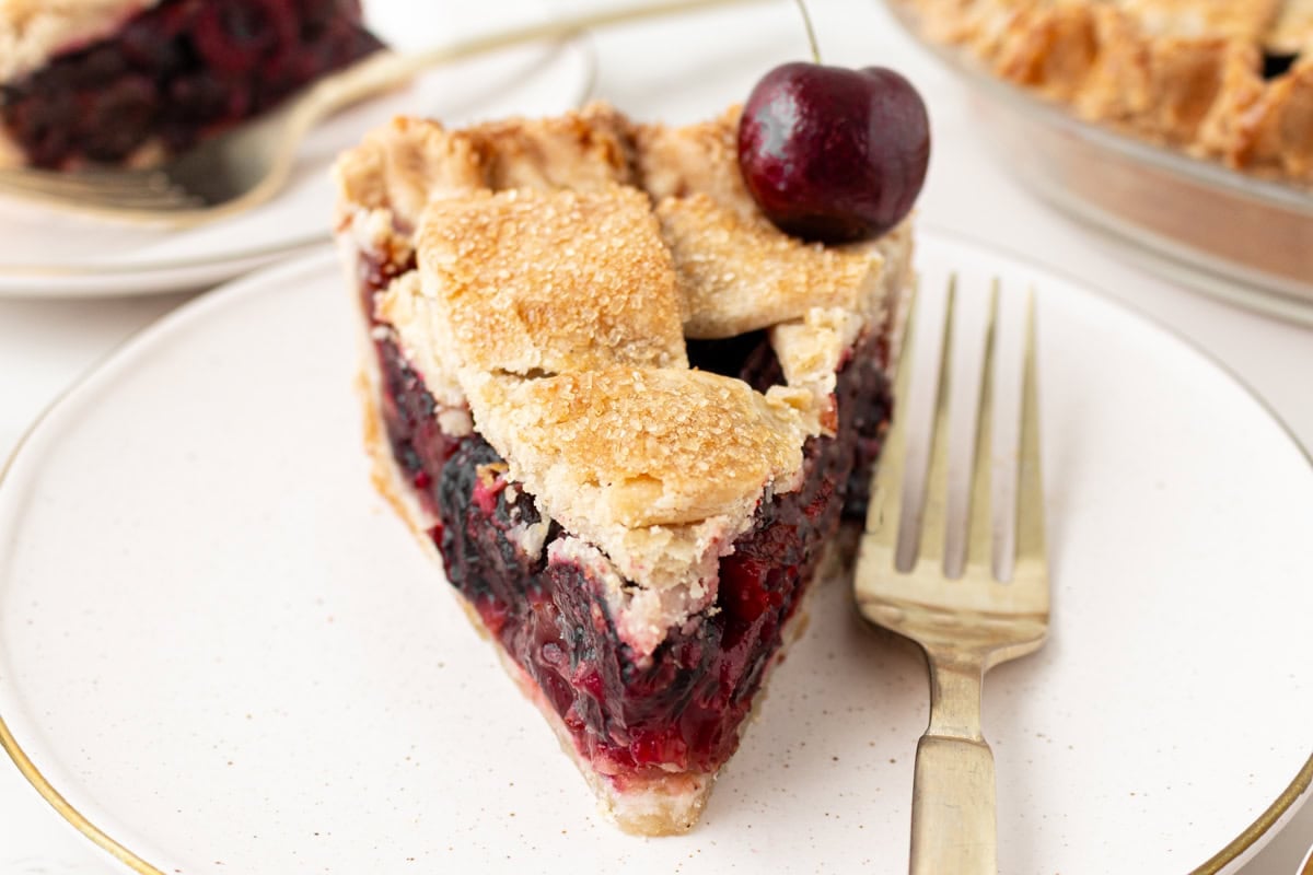 A slice of gluten-free cherry pie on a white plate with a gold rim, showing a golden sugar-crusted lattice top, a thick jammy cherry filling, and a fresh whole cherry placed on top, with a fork resting alongside and additional slices visible in the background.