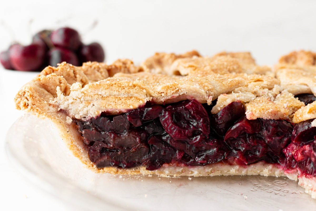 A close-up side view of a sliced gluten-free cherry pie on a white plate, showing the flaky golden crust and a thick, glossy deep red cherry filling packed with whole halved cherries, with fresh cherries blurred in the background.