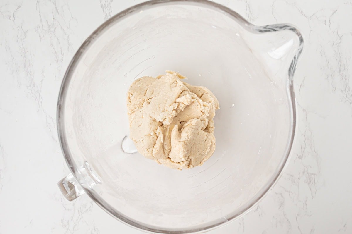 Overhead view of gluten-free pie crust dough formed into a rough ball in a glass stand mixer bowl on a white marble surface.