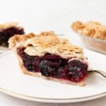 A close-up side view of a slice of gluten-free cherry pie on a white plate with a gold rim, showing a flaky golden lattice crust and a deep, jammy cherry filling, with the whole pie visible in the background.