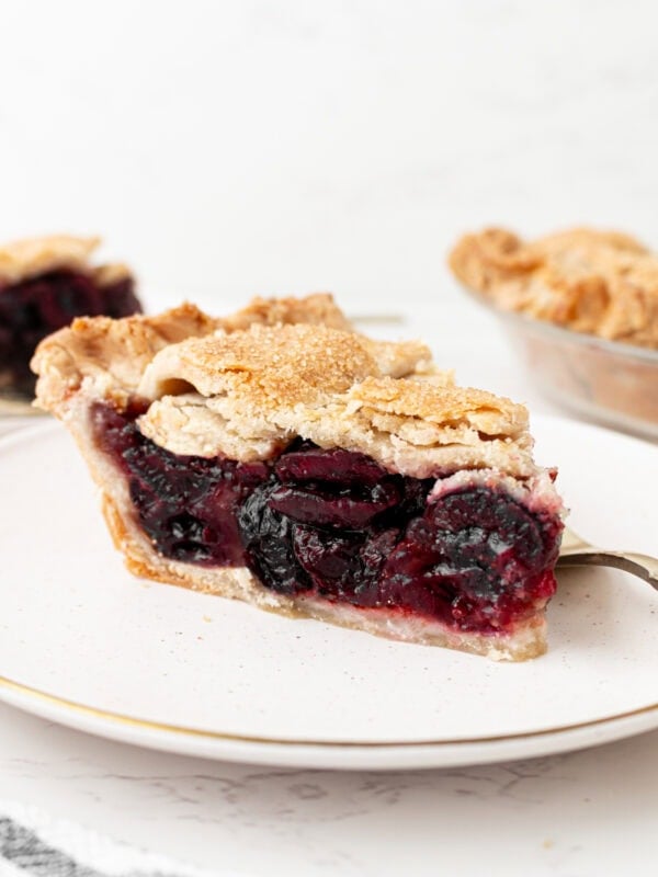 A close-up side view of a slice of gluten-free cherry pie on a white plate with a gold rim, showing a flaky golden lattice crust and a deep, jammy cherry filling, with the whole pie visible in the background.