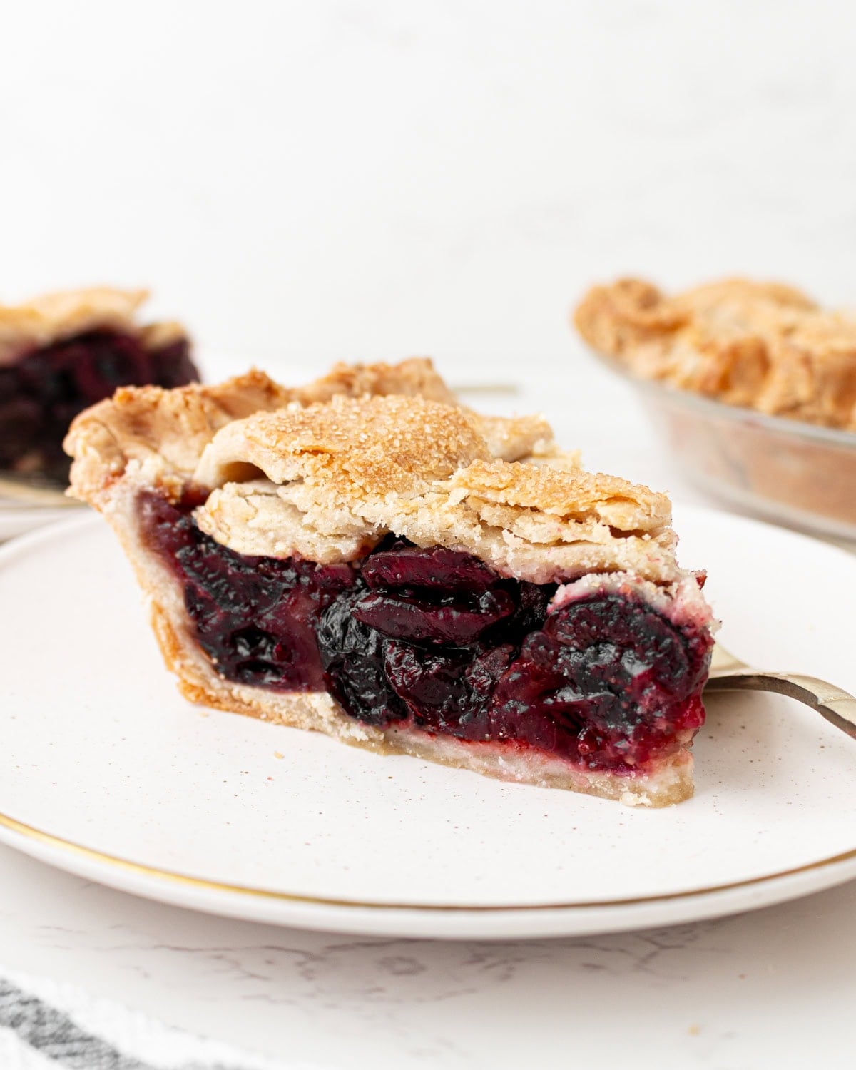 A close-up side view of a slice of gluten-free cherry pie on a white plate with a gold rim, showing a flaky golden lattice crust and a deep, jammy cherry filling, with the whole pie visible in the background.