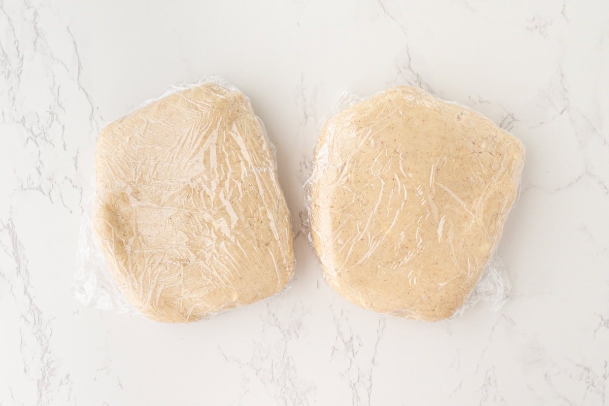 Two portions of gluten-free pie crust dough flattened into discs and wrapped tightly in plastic wrap, placed side by side on a white marble surface.
