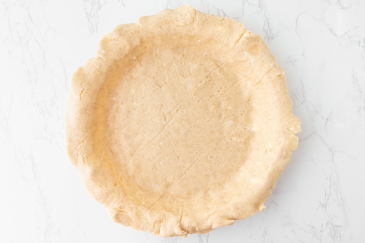 Overhead view of gluten-free pie crust dough fitted into a glass pie plate with the edges draped over the rim, on a white marble surface.