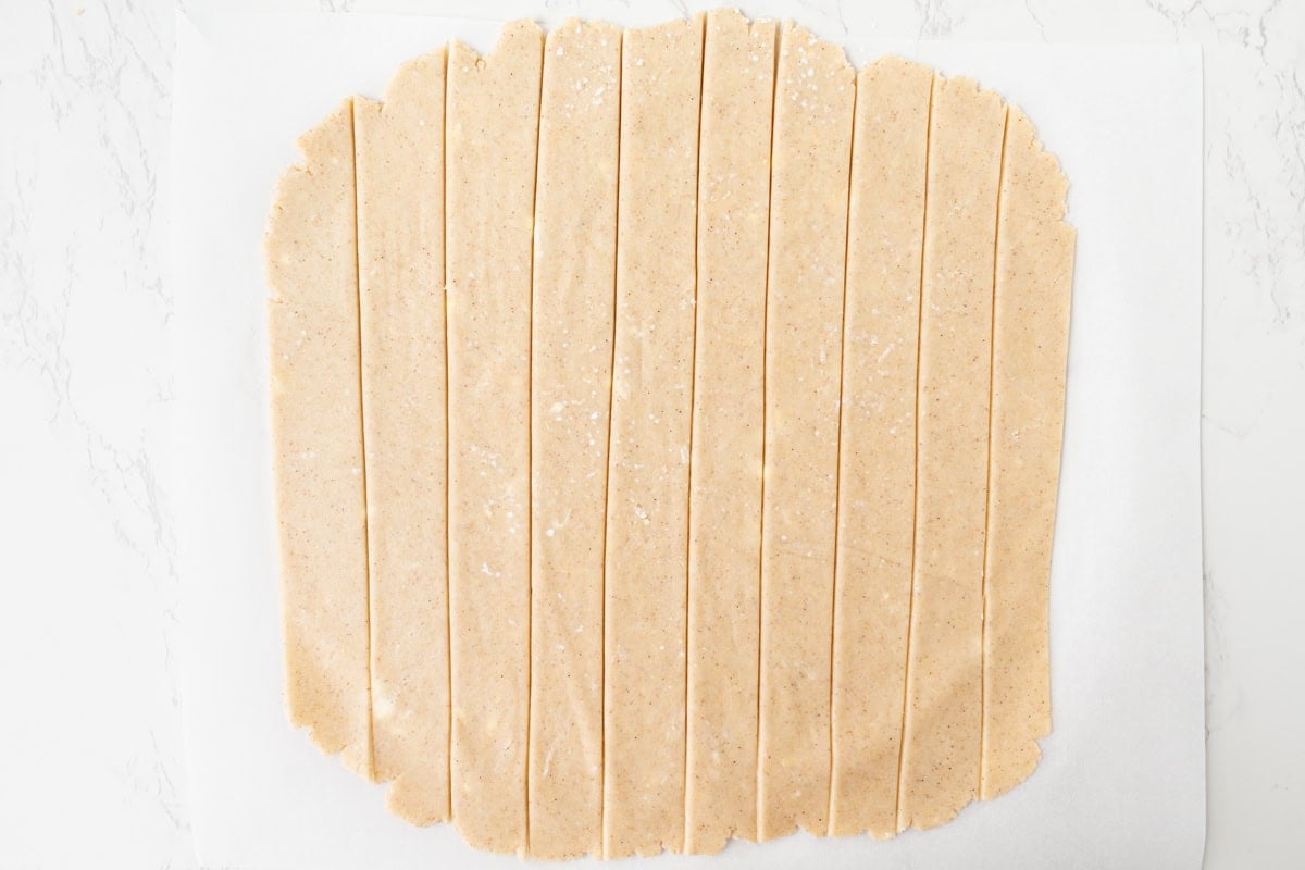 Overhead view of gluten-free pie crust dough rolled out and cut into even strips for a lattice top, laid out on parchment paper on a white marble surface.