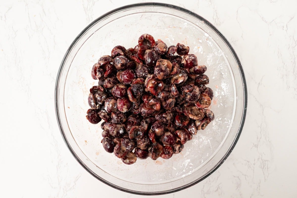 Overhead view of fresh pitted cherries tossed with sugar, cornstarch, and spices in a large glass mixing bowl on a white marble surface.
