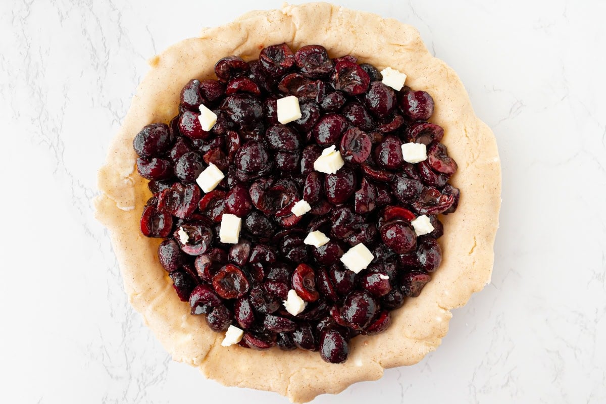 Overhead view of a gluten-free pie shell filled with glossy fresh cherry filling and dotted with small cubes of butter, ready for the lattice top.