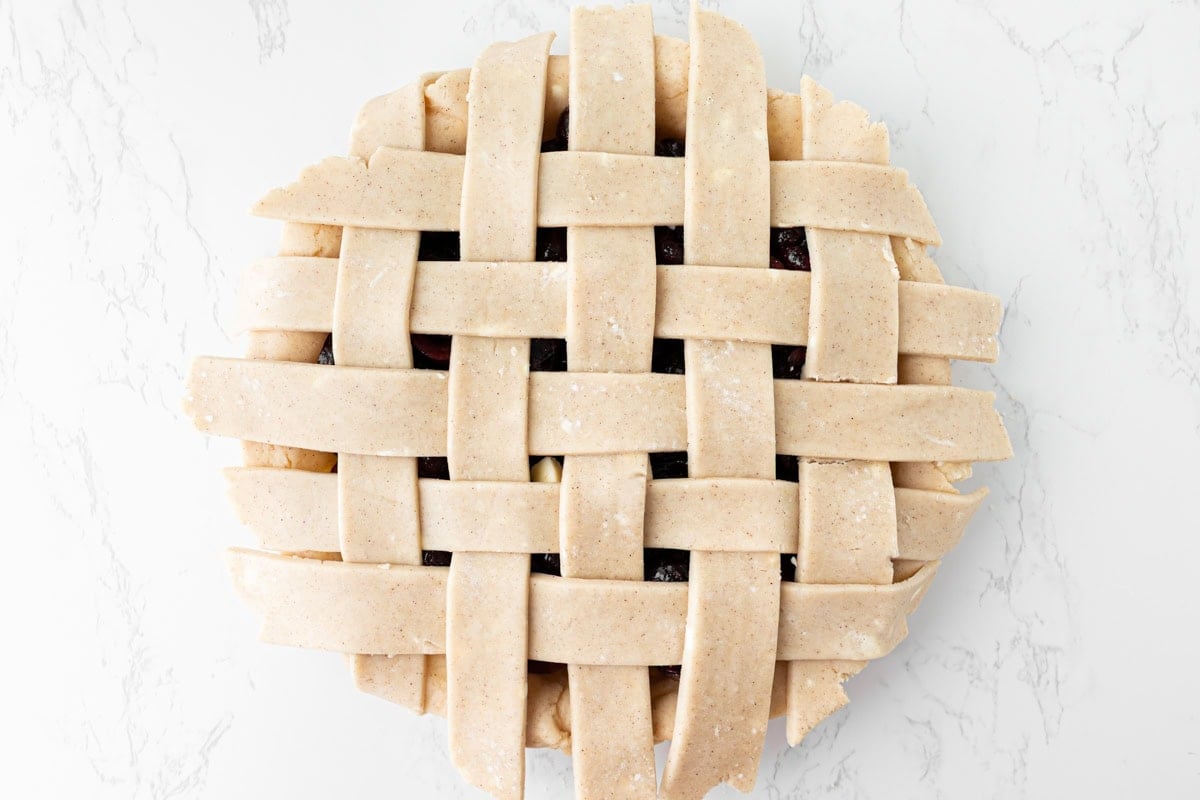Overhead view of a gluten-free cherry pie with a fully woven lattice crust before baking, showing the over-under strip pattern with the dark cherry filling visible through the gaps, on a white marble surface.