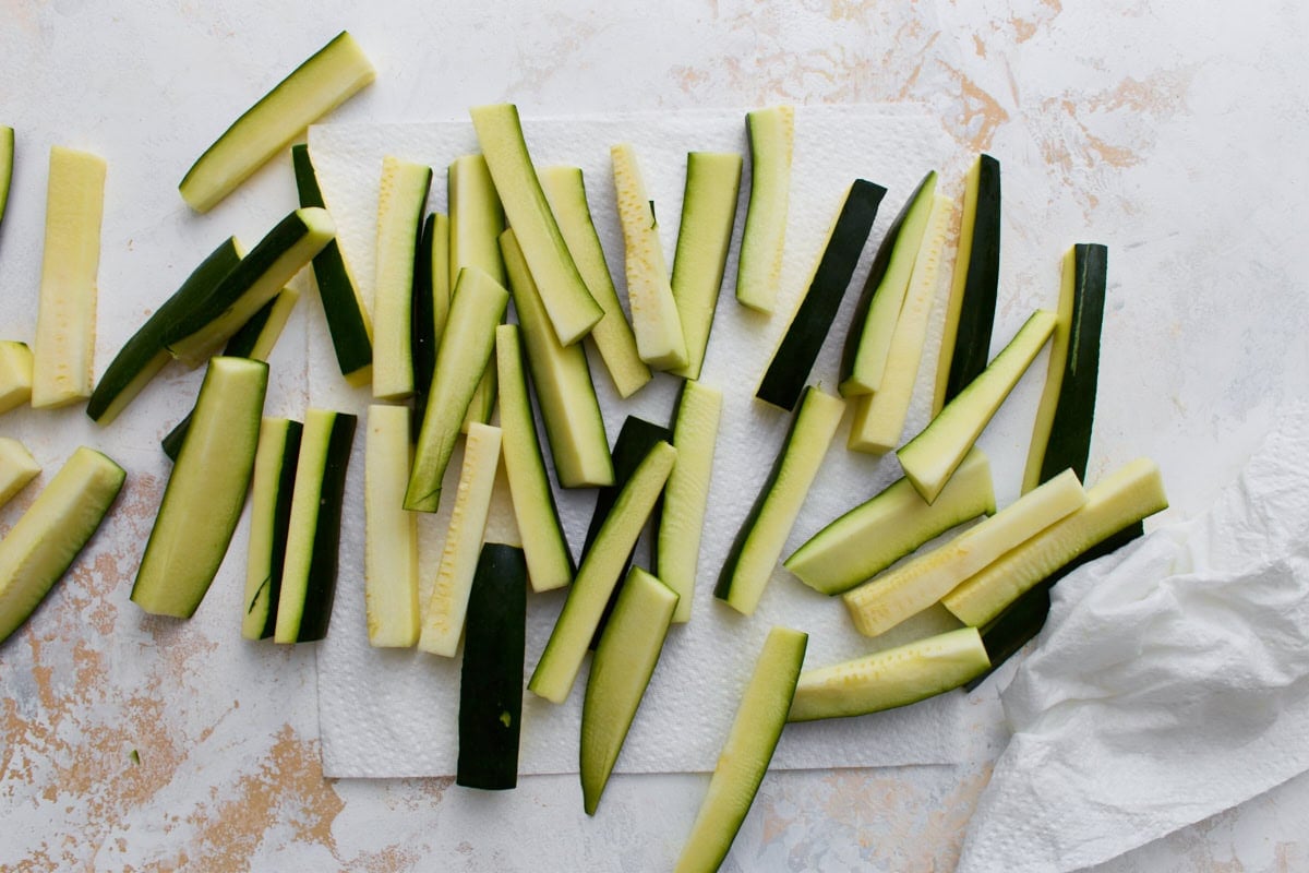 Fresh zucchini cut into fry-shaped sticks drying on paper towels before breading.