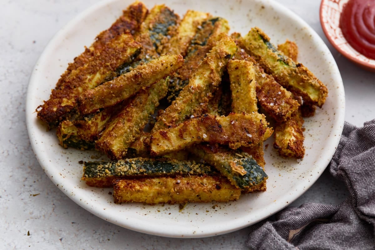 Crispy baked keto zucchini fries piled on a white plate with flaky salt and a side of ketchup for dipping.