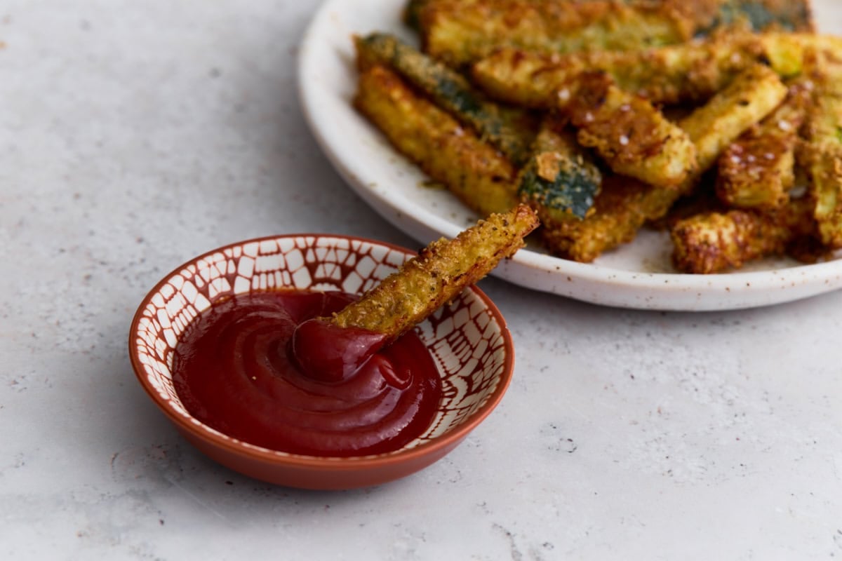 Parmesan-crusted zucchini fry dipped into ketchup with a plate of crispy baked zucchini fries in the background.