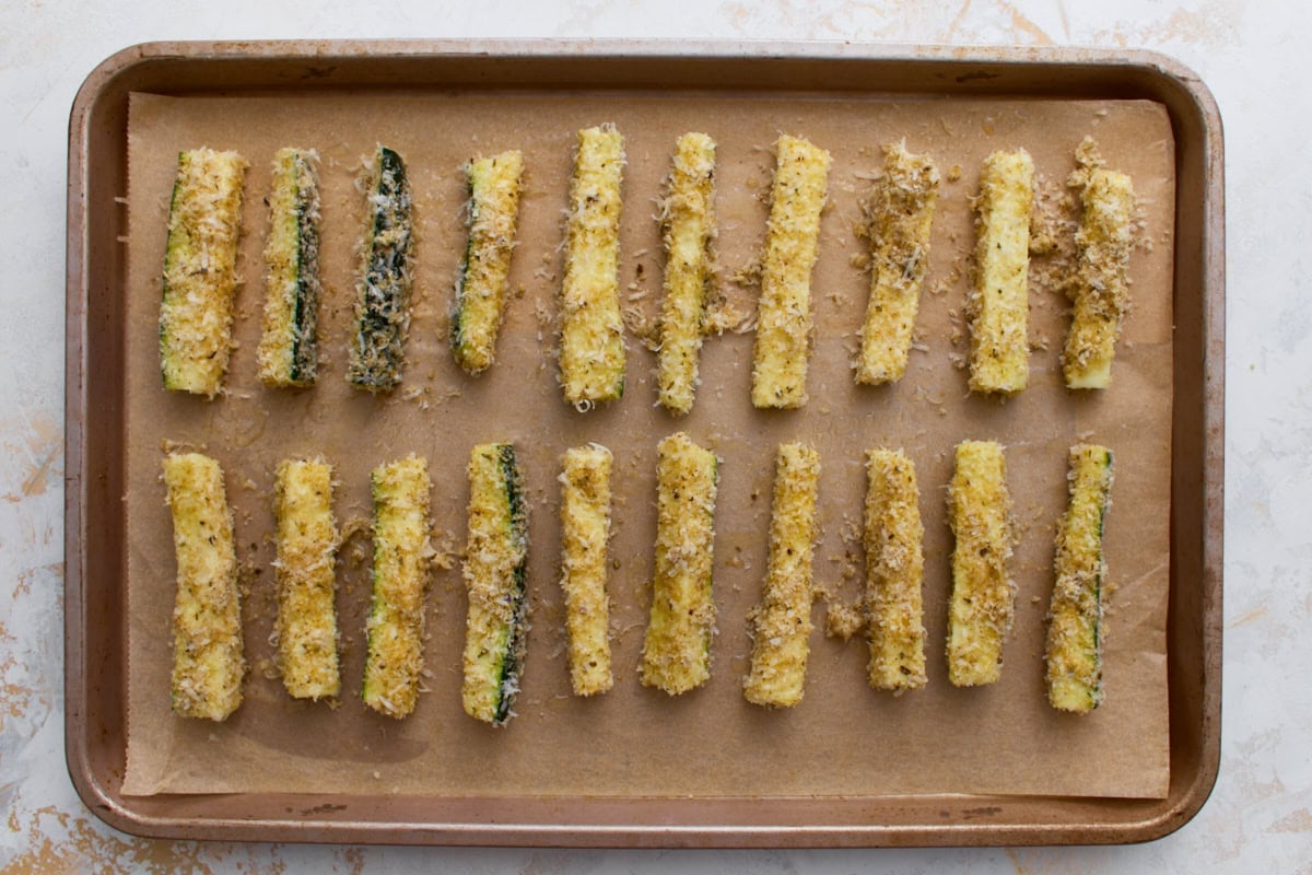 Breaded zucchini fries arranged in a single layer on a parchment-lined baking sheet before baking.