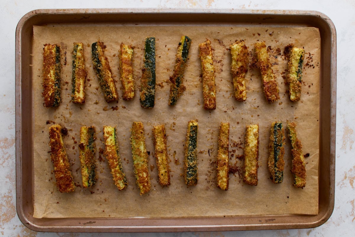 Golden brown baked keto zucchini fries on a parchment-lined baking sheet after roasting until crispy.