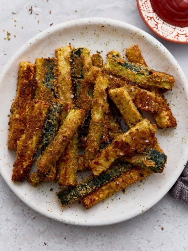 Crispy baked keto zucchini fries coated in Parmesan and almond flour served on a white plate with a side of ketchup.