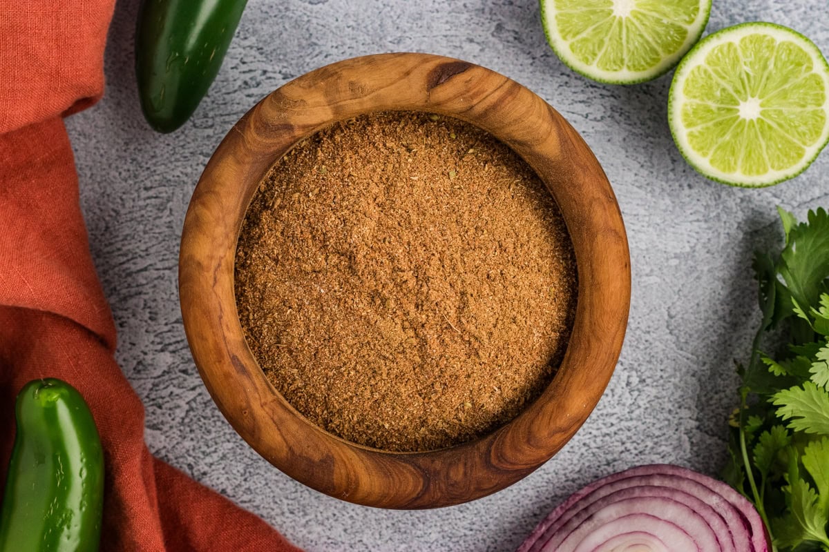 overhead view of homemade gluten-free taco seasoning in a wooden bowl surrounded by jalapeños, lime, cilantro, and red onion