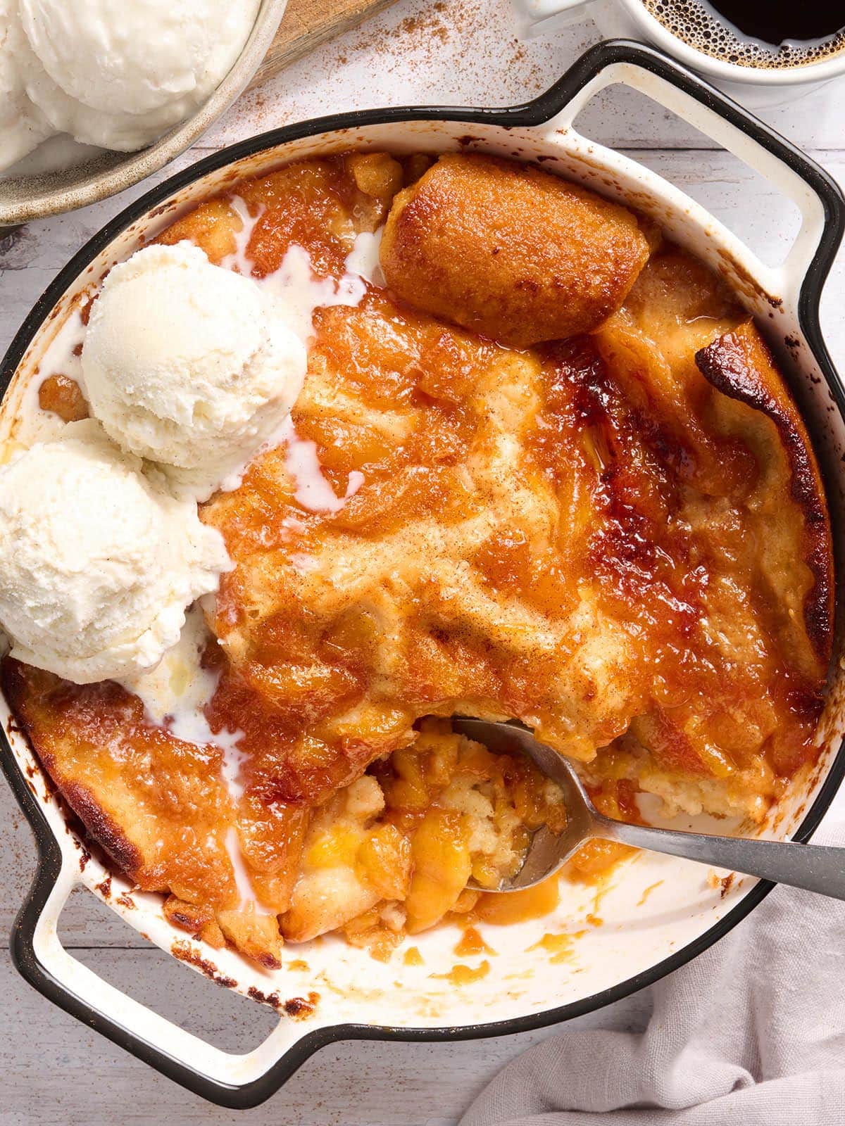 Gluten free peach cobbler in a white baking dish with three scoops of vanilla ice cream melting into the golden peach filling
