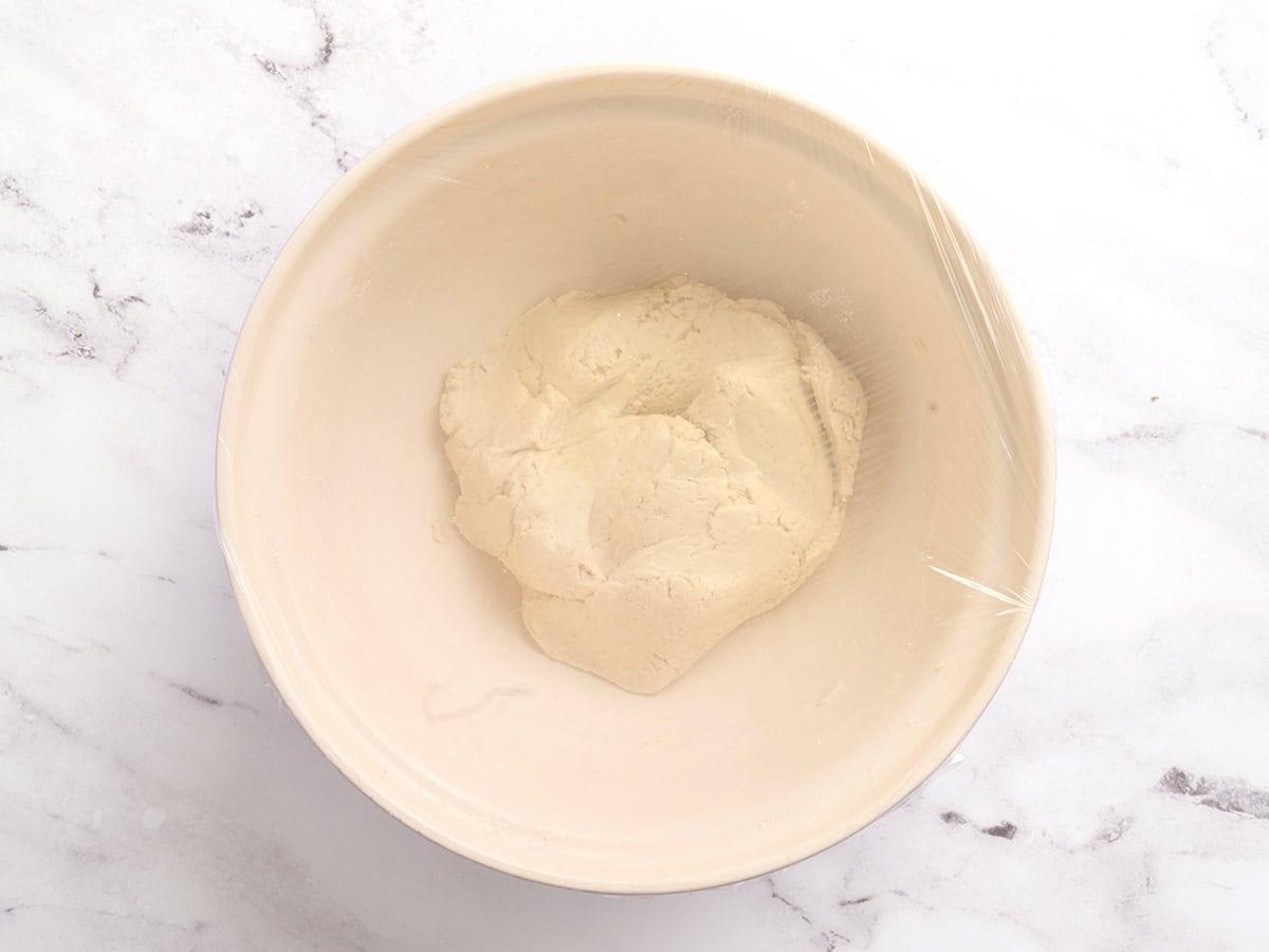 Soft, sticky gluten free pita dough in a bowl covered with plastic wrap, ready to rise.