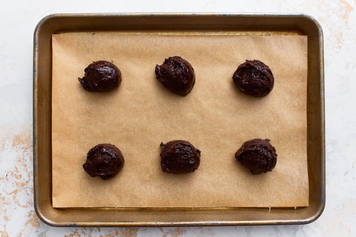 Six scoops of flourless chocolate cookie dough spaced on a parchment-lined baking sheet, ready to bake.