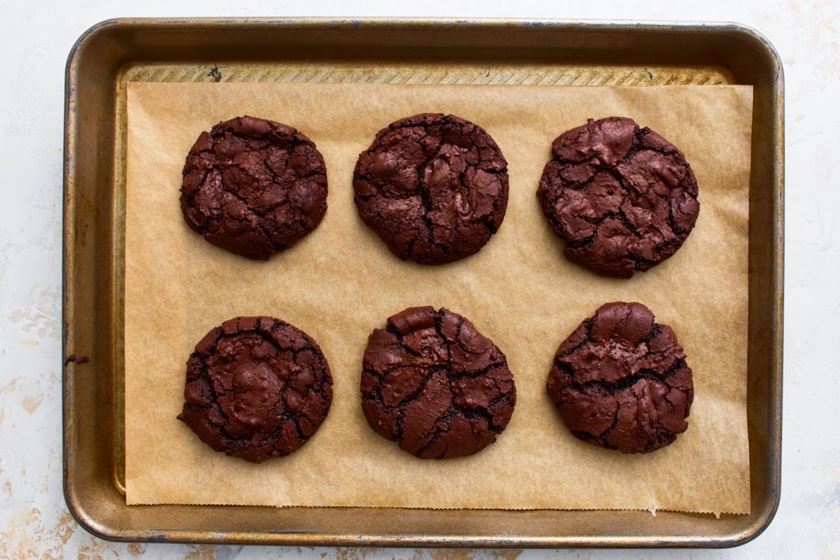 Six flourless chocolate cookies fresh out of the oven with shiny, cracked tops.