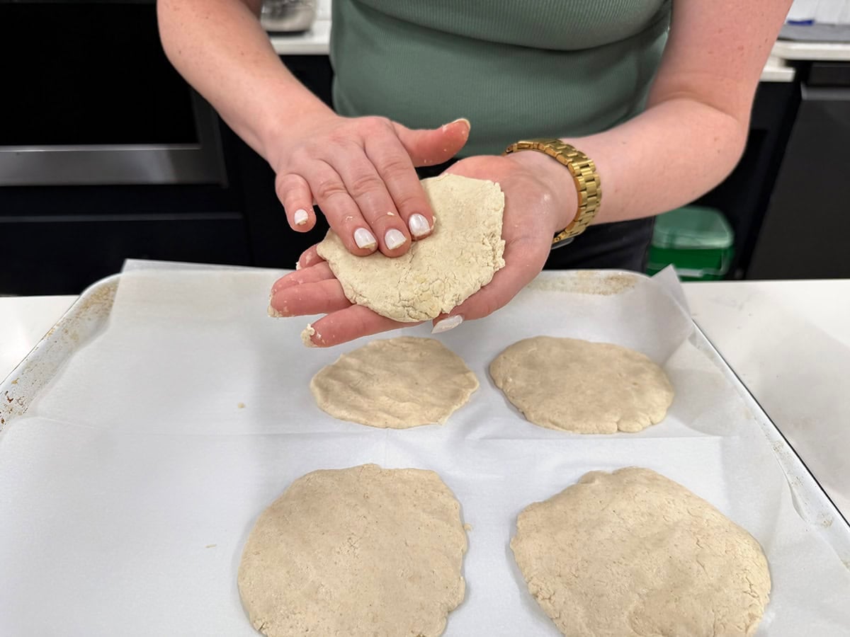 Pressing gluten free pita dough into a round with oiled hands over a parchment-lined sheet pan.