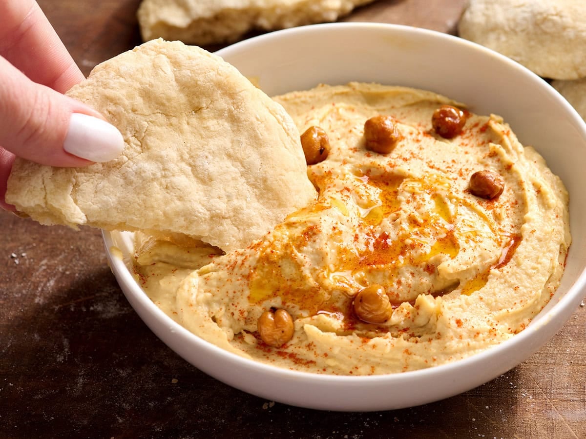 Gluten free pita bread being dipped into a bowl of hummus topped with chickpeas and paprika.