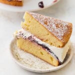 Slice of gluten-free Victoria sponge cake on a speckled plate, showing layers of sponge, jam, and whipped cream dusted with powdered sugar