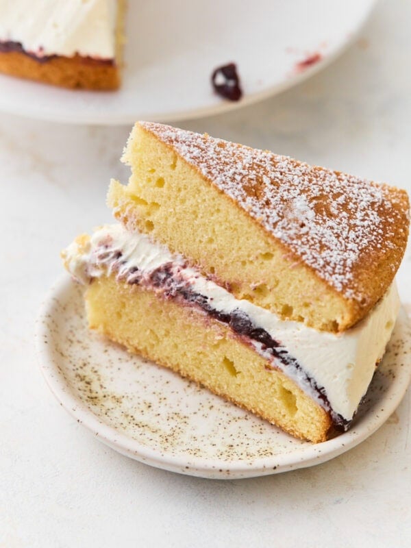 Slice of gluten-free Victoria sponge cake on a speckled plate, showing layers of sponge, jam, and whipped cream dusted with powdered sugar