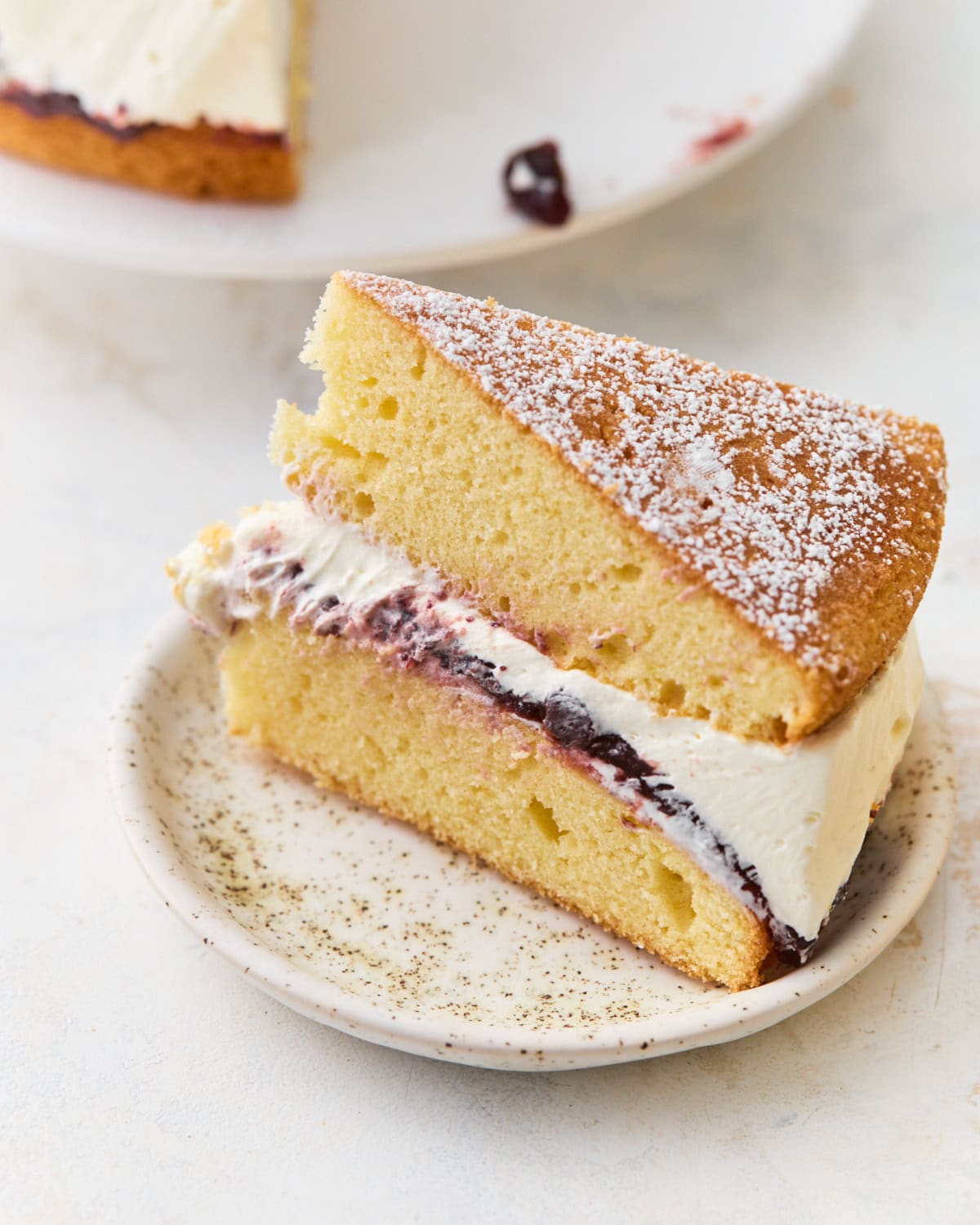 Slice of gluten-free Victoria sponge cake on a speckled plate, showing layers of sponge, jam, and whipped cream dusted with powdered sugar