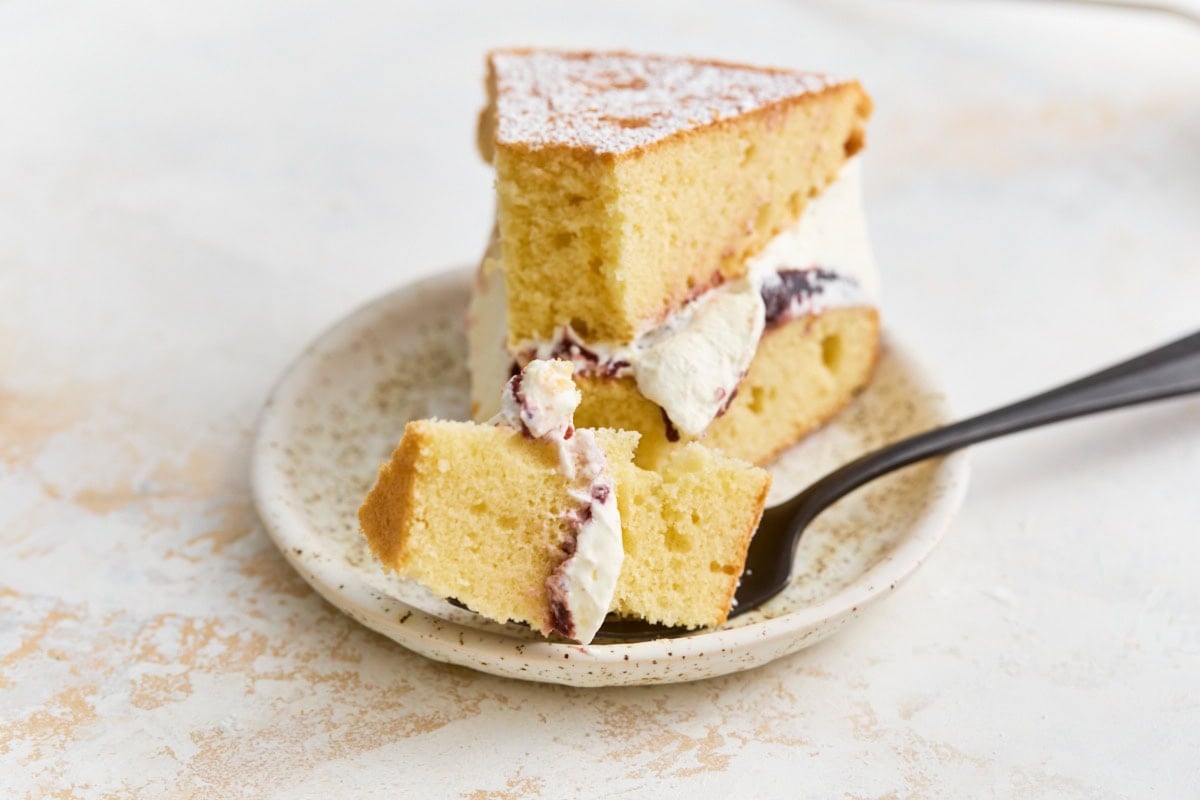 Slice of gluten-free Victoria sponge cake on a plate with a bite on a fork showing the soft crumb