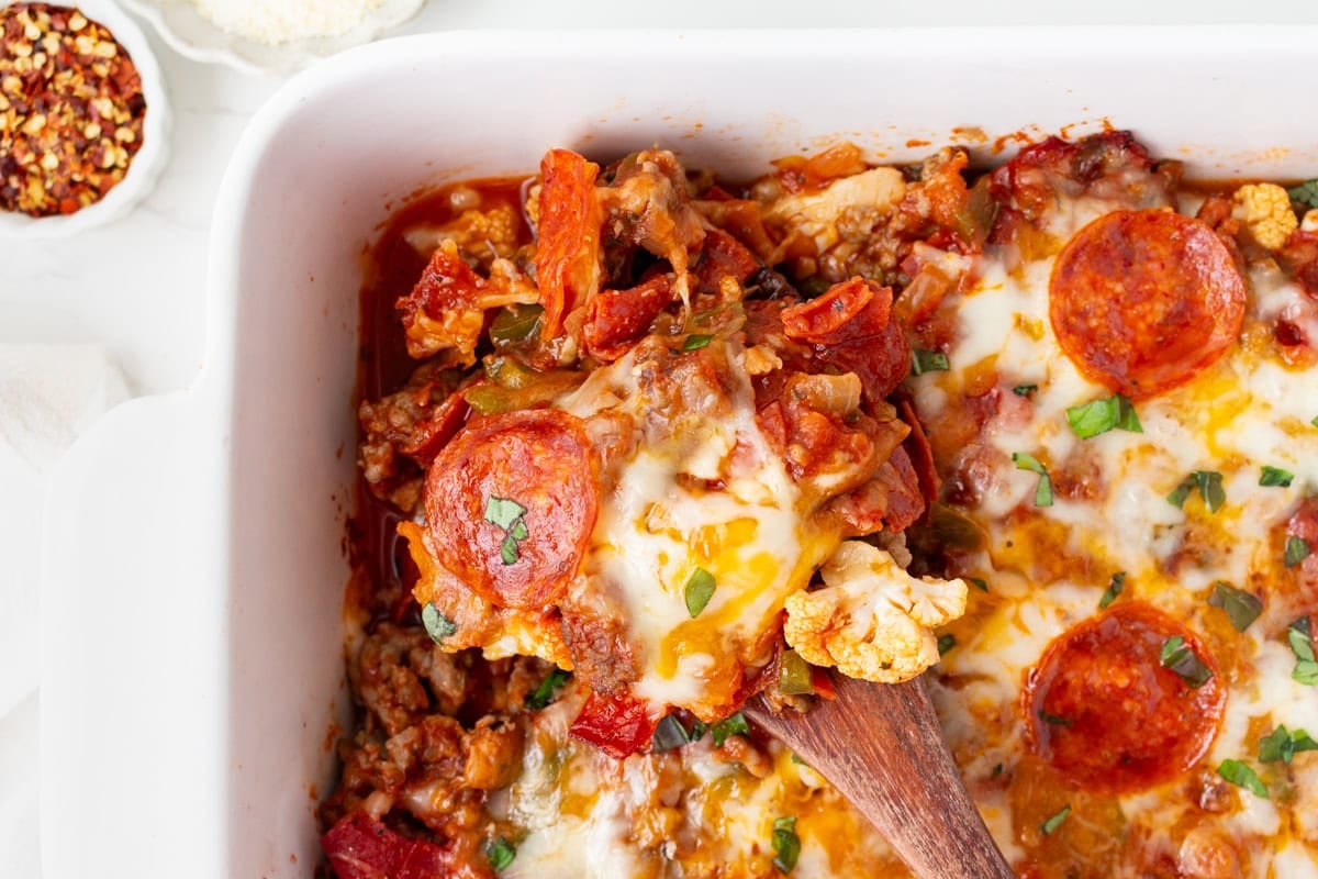 Close-up of keto pizza casserole in the baking dish with a wooden spoon lifting out a cheesy serving.
