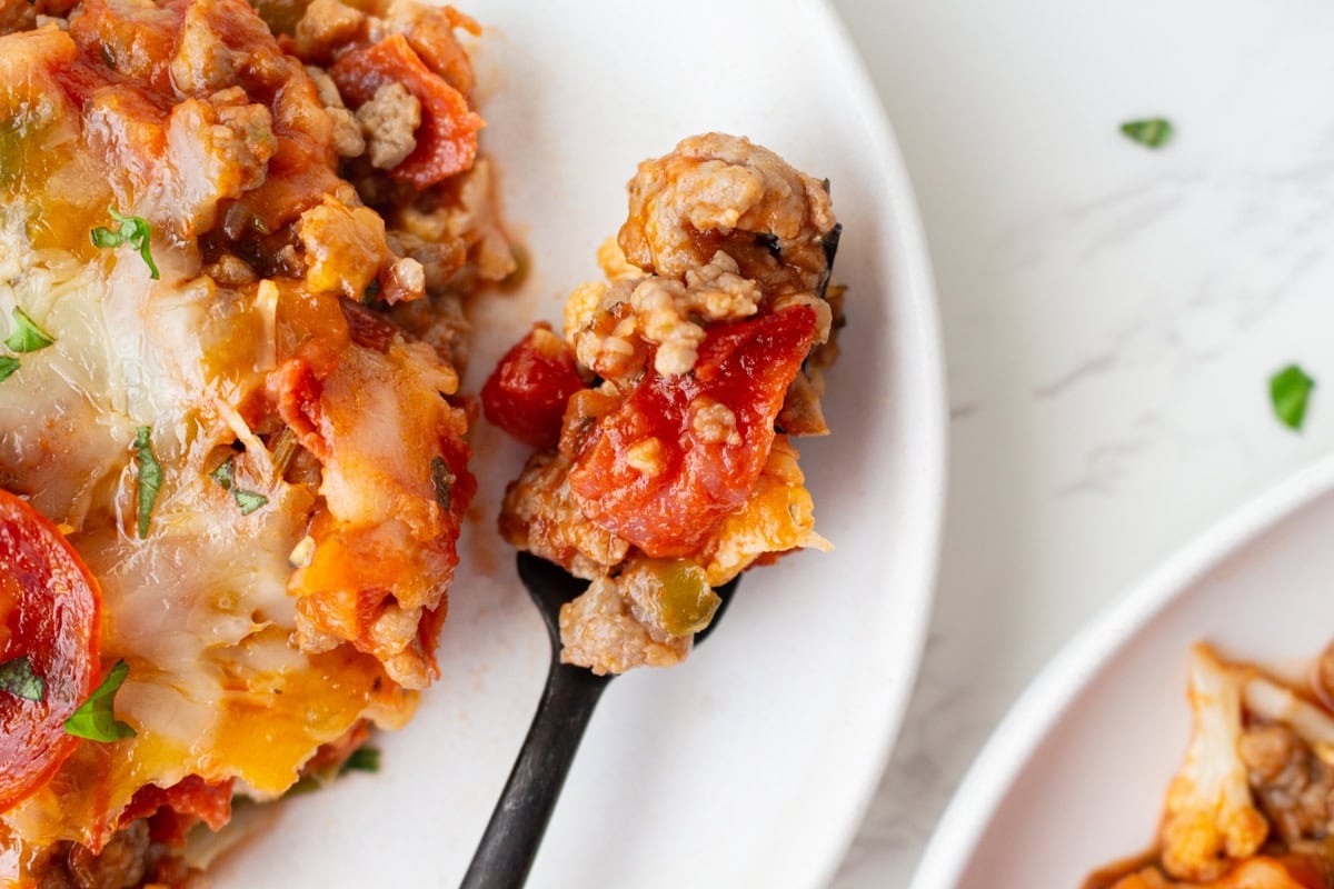 Close-up of a spoonful of keto pizza casserole beside two plated servings.