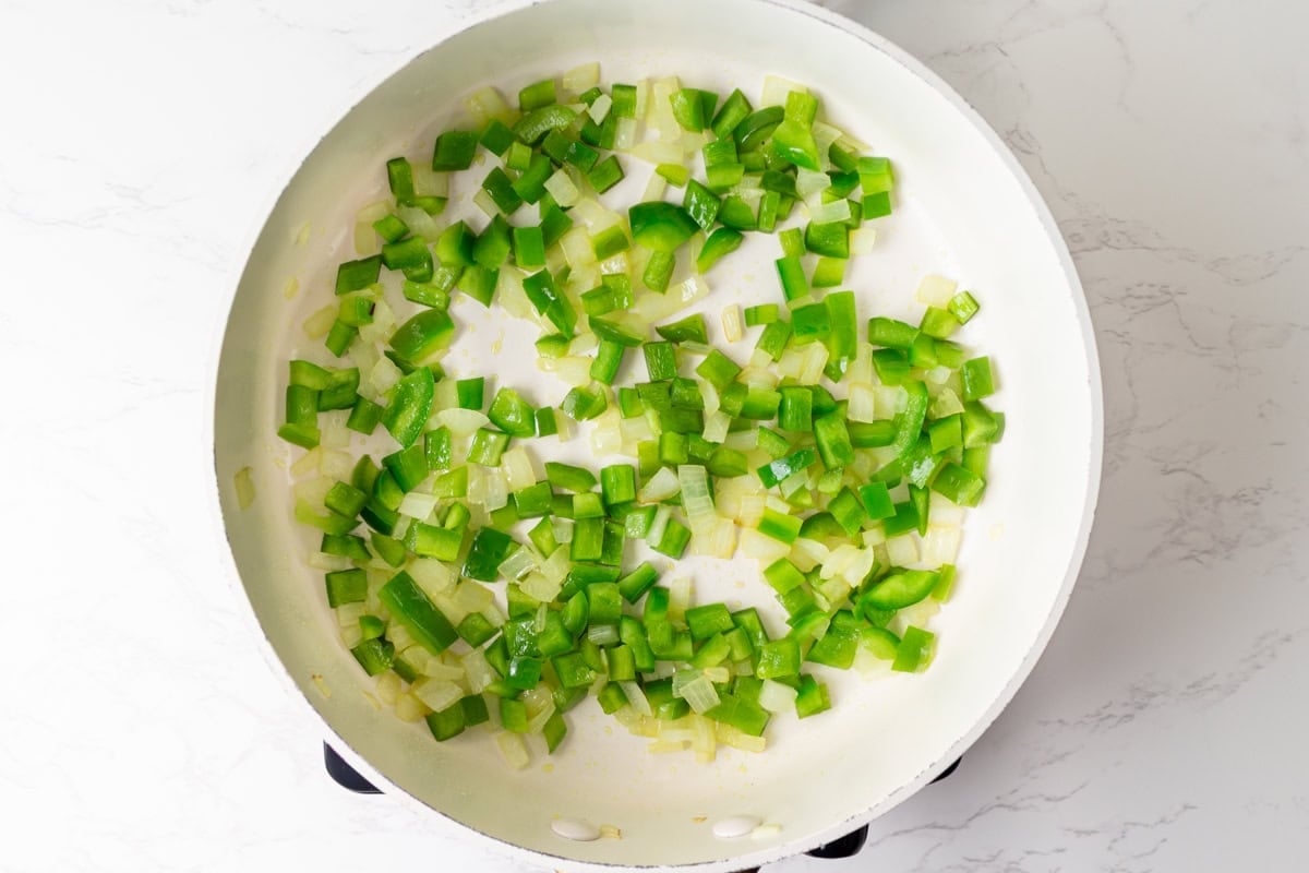 Diced onion and green bell pepper sautéing in a white skillet.