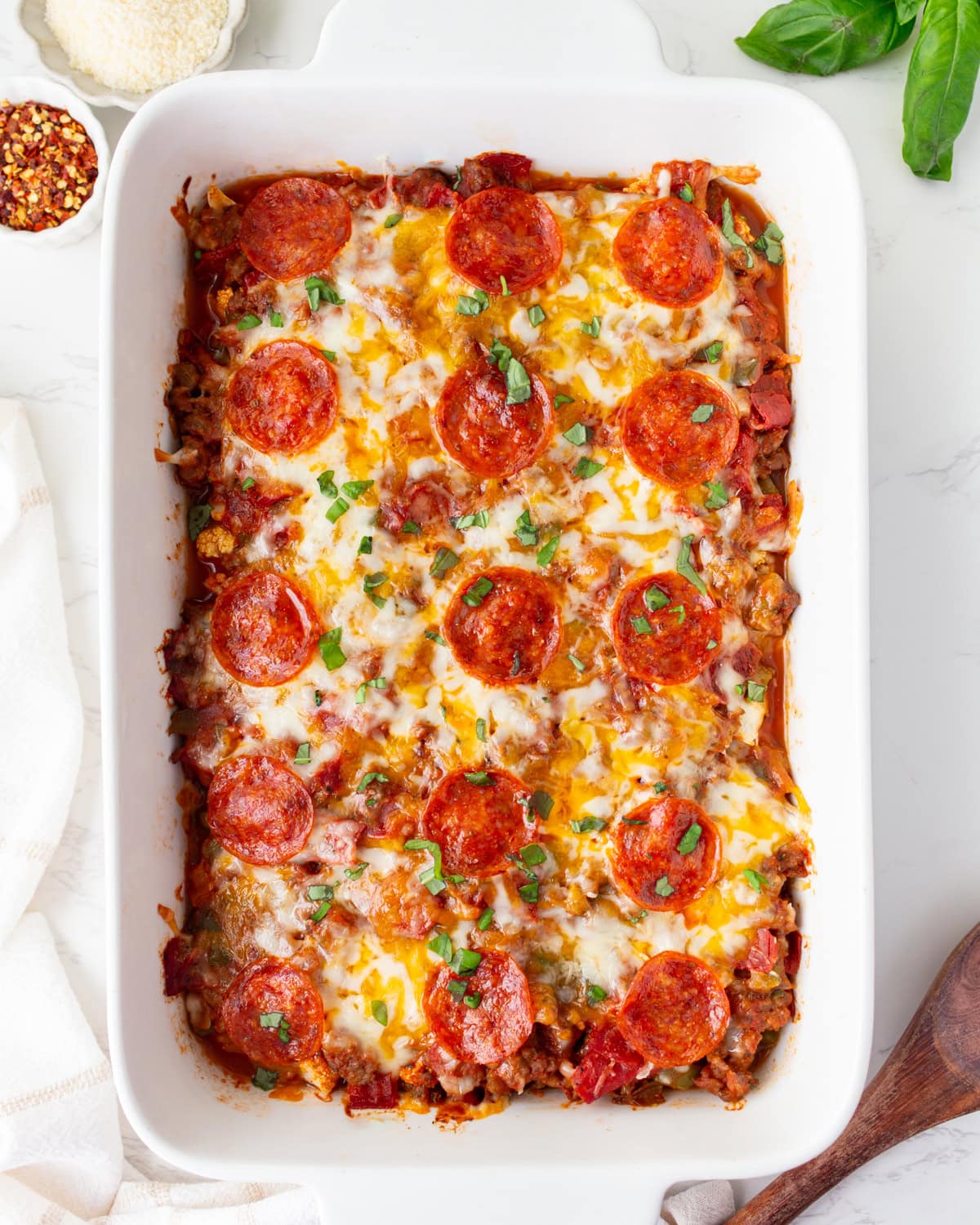 Overhead shot of keto pizza casserole in a white baking dish, topped with melted cheese, pepperoni, and fresh basil.