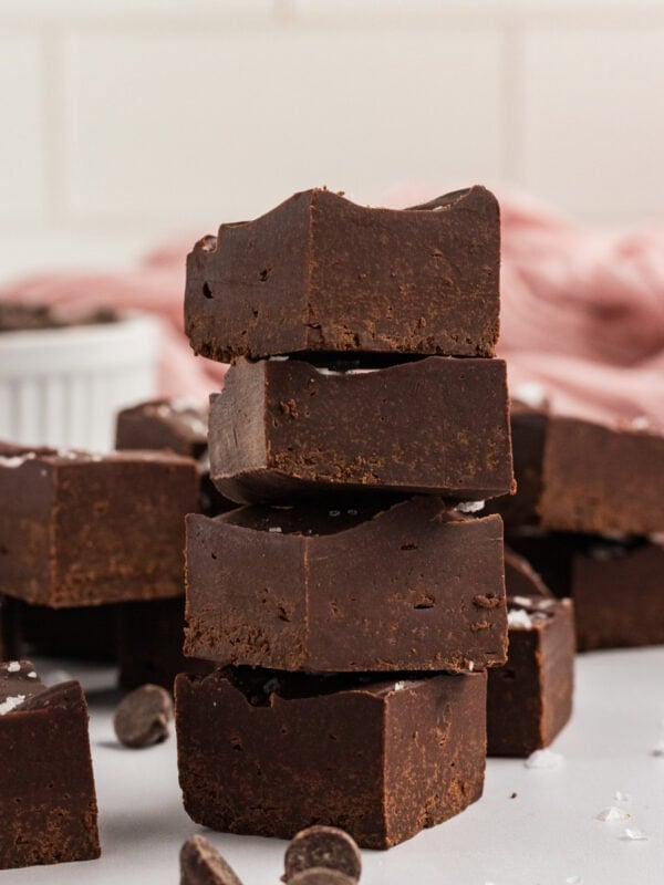Stack of gluten-free dairy-free chocolate fudge squares topped with flaky sea salt, with chocolate chips scattered around and a pink cloth in the background