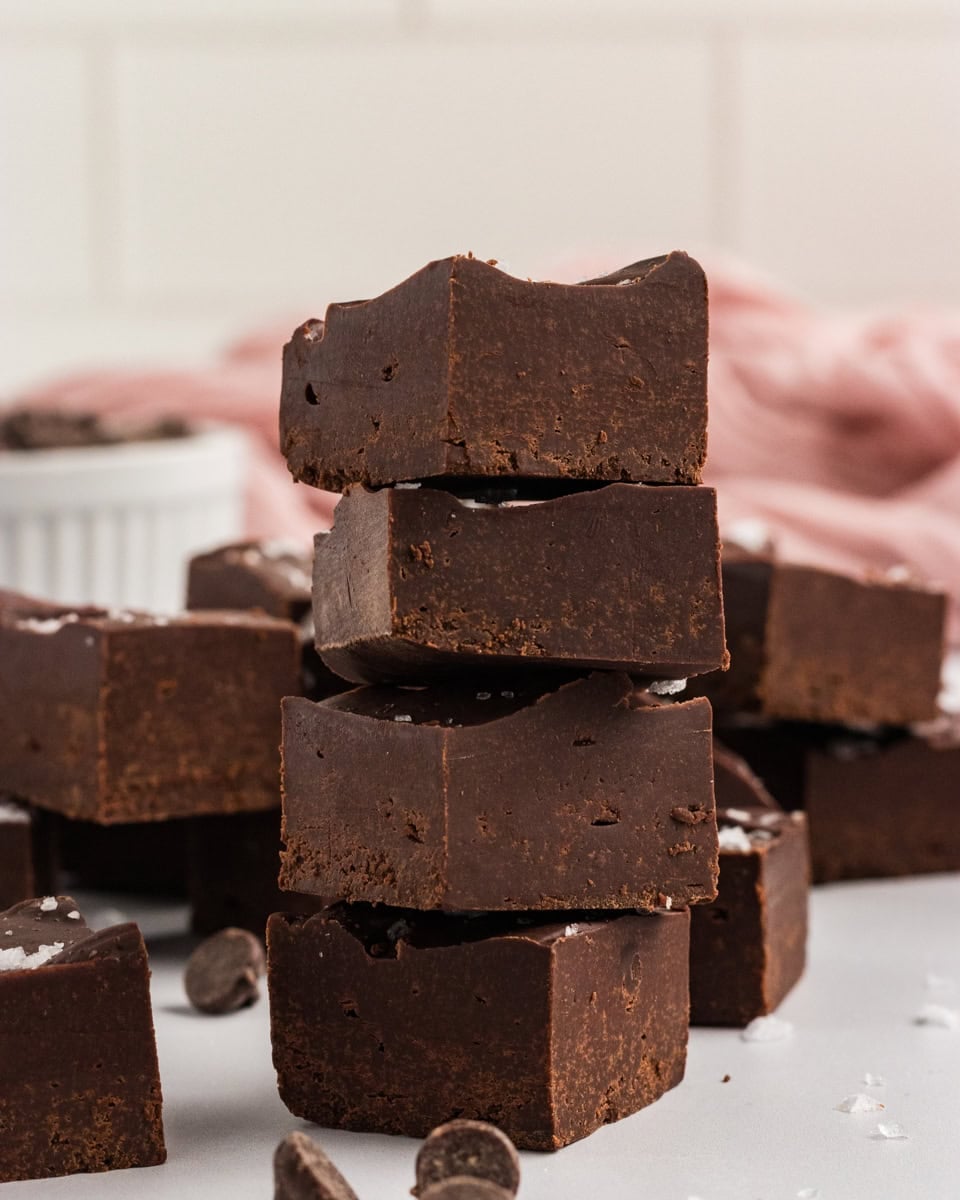 Stack of gluten-free dairy-free chocolate fudge squares topped with flaky sea salt, with chocolate chips scattered around and a pink cloth in the background