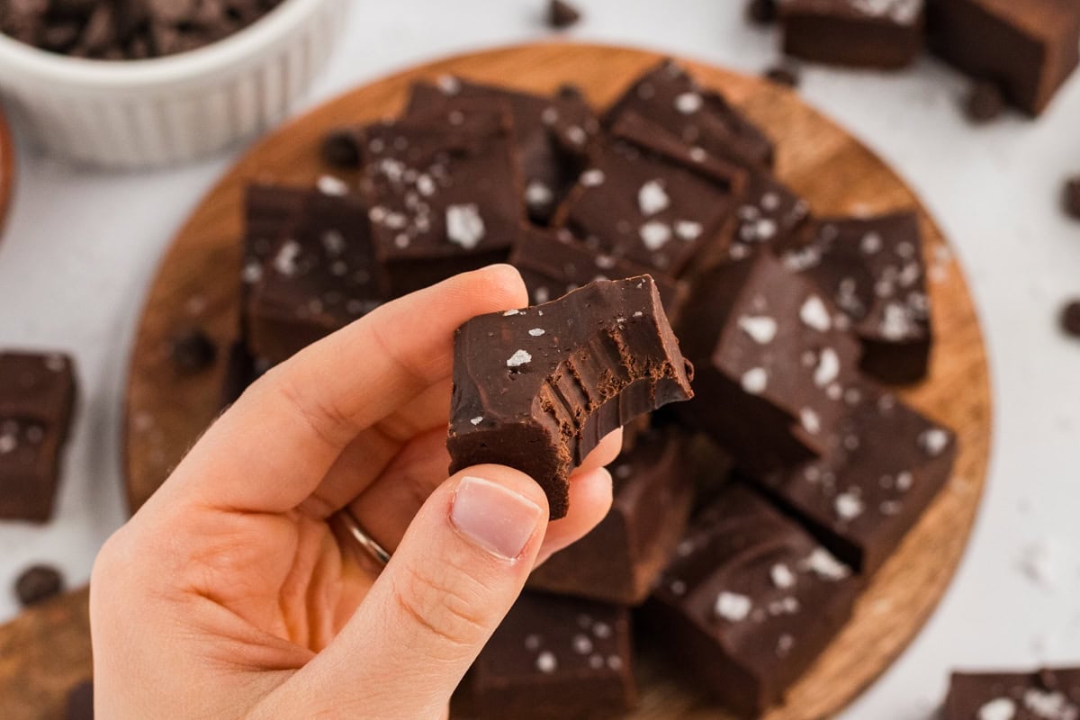 Hand holding a bitten square of chocolate fudge, showing the dense, fudgy texture and flaky salt topping